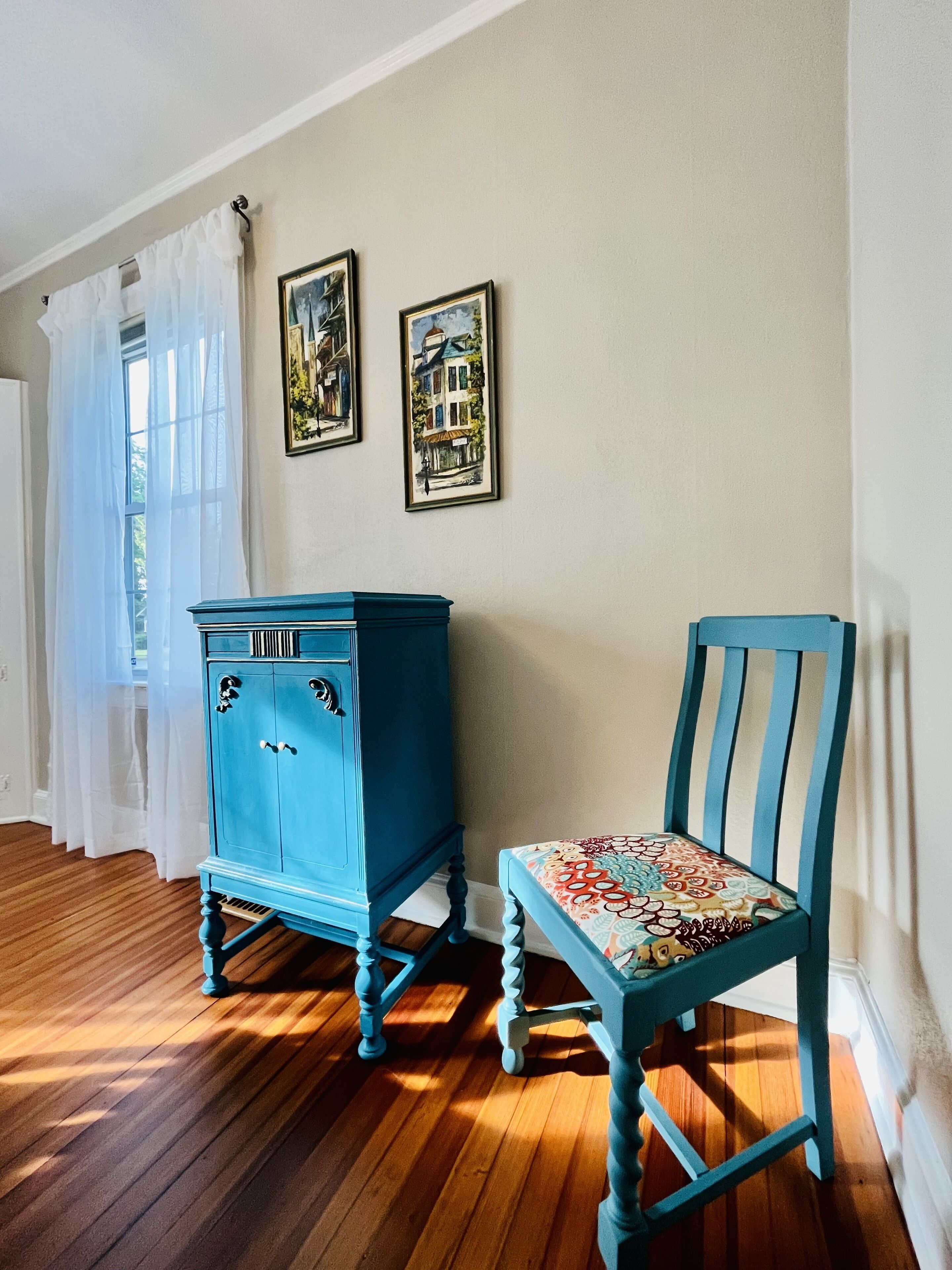 Quarto com piso de madeira, um armário azul, uma cadeira azul com almofada estampada e duas molduras na parede.