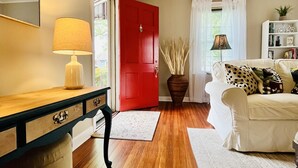 A living room with a wooden floor, a white sofa with patterned cushions, a red door, and a lamp on a wooden console table. - Historic Cozy Parkside Cottage in Savannah (Savannah)