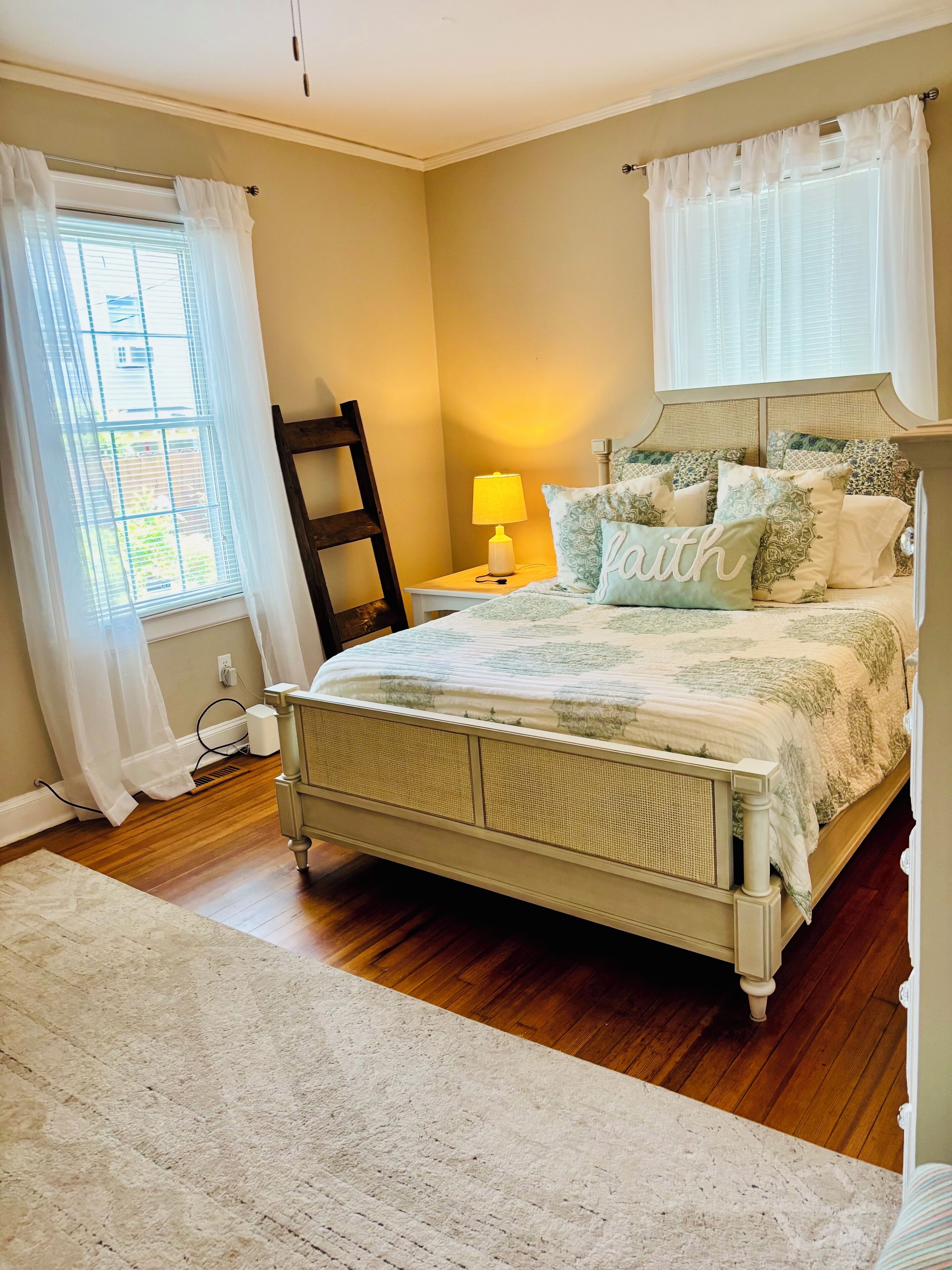 A bedroom with a bed, a bedside table with a lamp, a window with curtains, and a wooden floor.