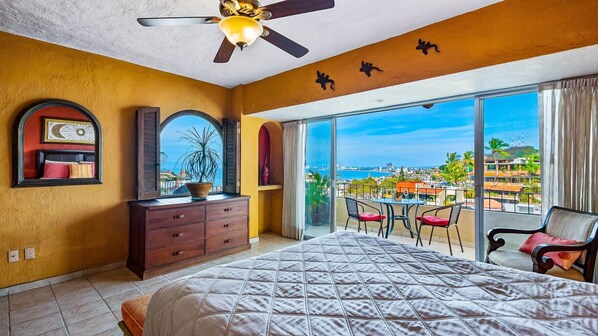 A hotel room with a large bed, a dresser, a ceiling fan, and a balcony with a view of the city and sea.