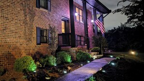 A brick house with a front porch, an American flag, and a well-lit garden.