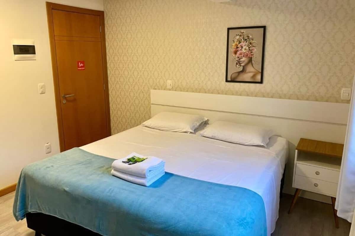 A hotel room with a bed, a nightstand, a wall-mounted artwork, and a door with a red sign.