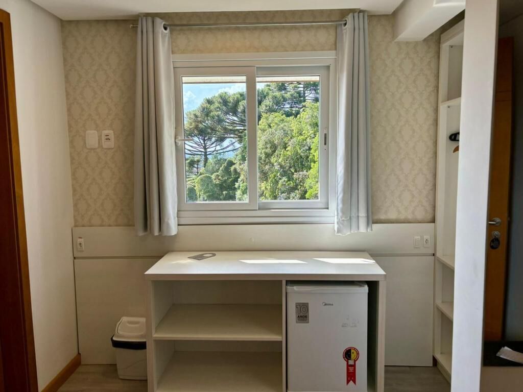 Una habitación pequeña con ventana, un escritorio blanco, un refrigerador pequeño y un cesto de basura.