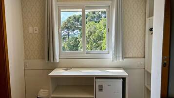 Una habitación pequeña con ventana, un escritorio blanco, un refrigerador pequeño y un cesto de basura.