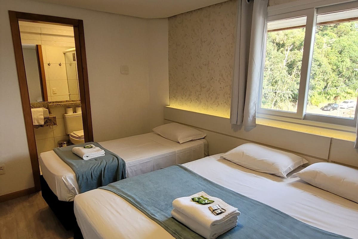 A hotel room with three beds, a bathroom visible through an open door, and a window with a view of greenery.