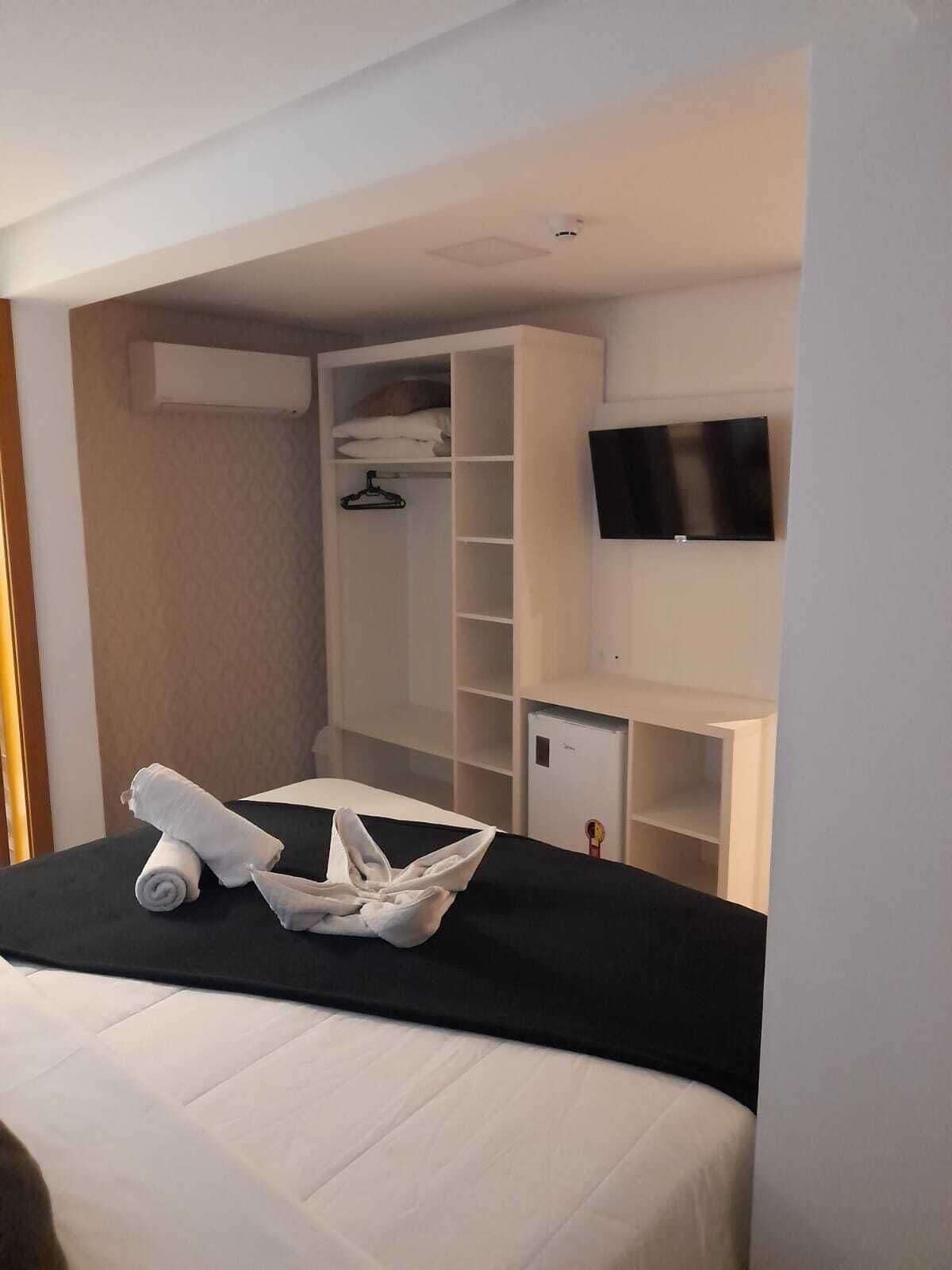 A hotel room with a bed, a TV mounted on the wall, a wardrobe, and a small desk with a chair.