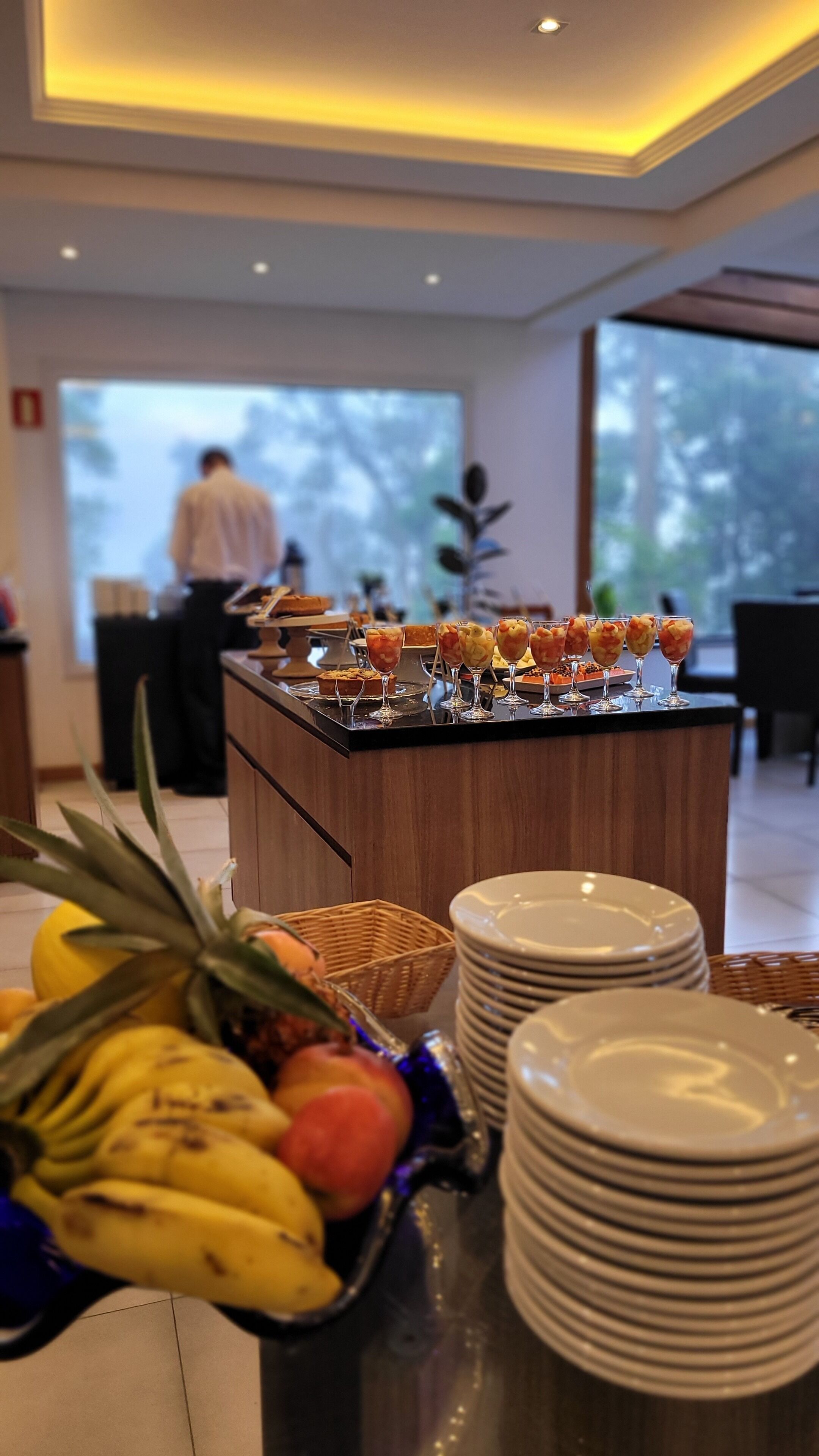 Disposição de buffet com frutas e bebidas, uma pilha de pratos e uma vista de uma pessoa ao fundo.