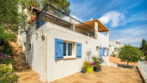 Un bâtiment blanc avec des volets bleus, un balcon doté d’une balustrade en bois et une terrasse carrelée agrémentée de plantes en pot.