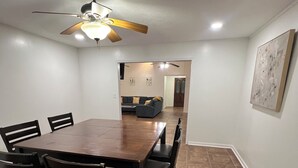 A dining area with a wooden table and chairs, a ceiling fan, and a view into a living room with a couch and a door. - The Simple Dwelling Place (Sulphur)