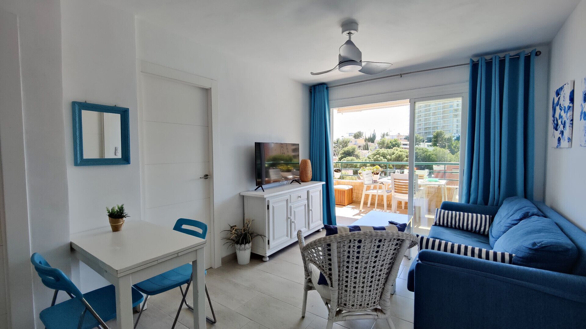 Un salón luminoso con una puerta blanca, una mesa de comedor con sillas azules, un sofá azul y vistas a zonas verdes al exterior.