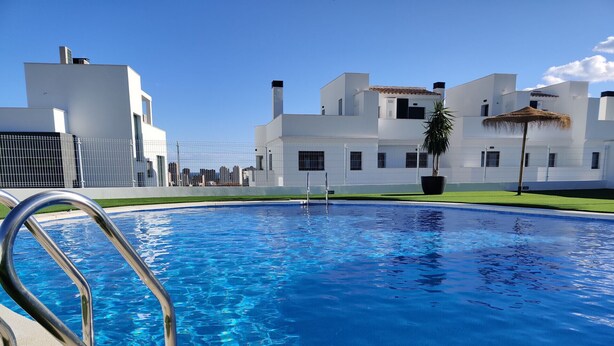 Une piscine entourée de rampes métalliques, bordée d’un bâtiment blanc doté de nombreuses fenêtres, sous un ciel d’un bleu limpide.
