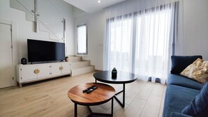 Un salon moderne avec un téléviseur à écran plat posé sur un meuble blanc, une table basse ronde avec une télécommande, un canapé bleu agrémenté d’un coussin décoratif et un escalier menant à l’étage supérieur.