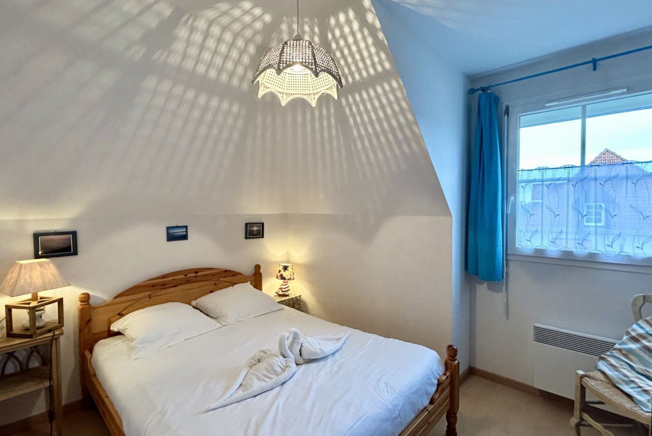Une chambre à coucher comprenant un lit, une table de chevet, une lampe, une fenêtre avec des rideaux et un luminaire au plafond.