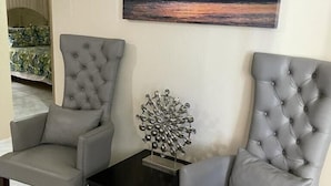 Two grey tufted armchairs facing each other with a small table between them, a metal sculpture on the table, and a sunset painting on the wall.