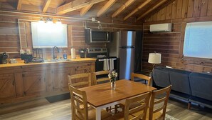 A rustic cabin interior with a wooden dining table, chairs, a kitchen area with appliances, and a sofa.