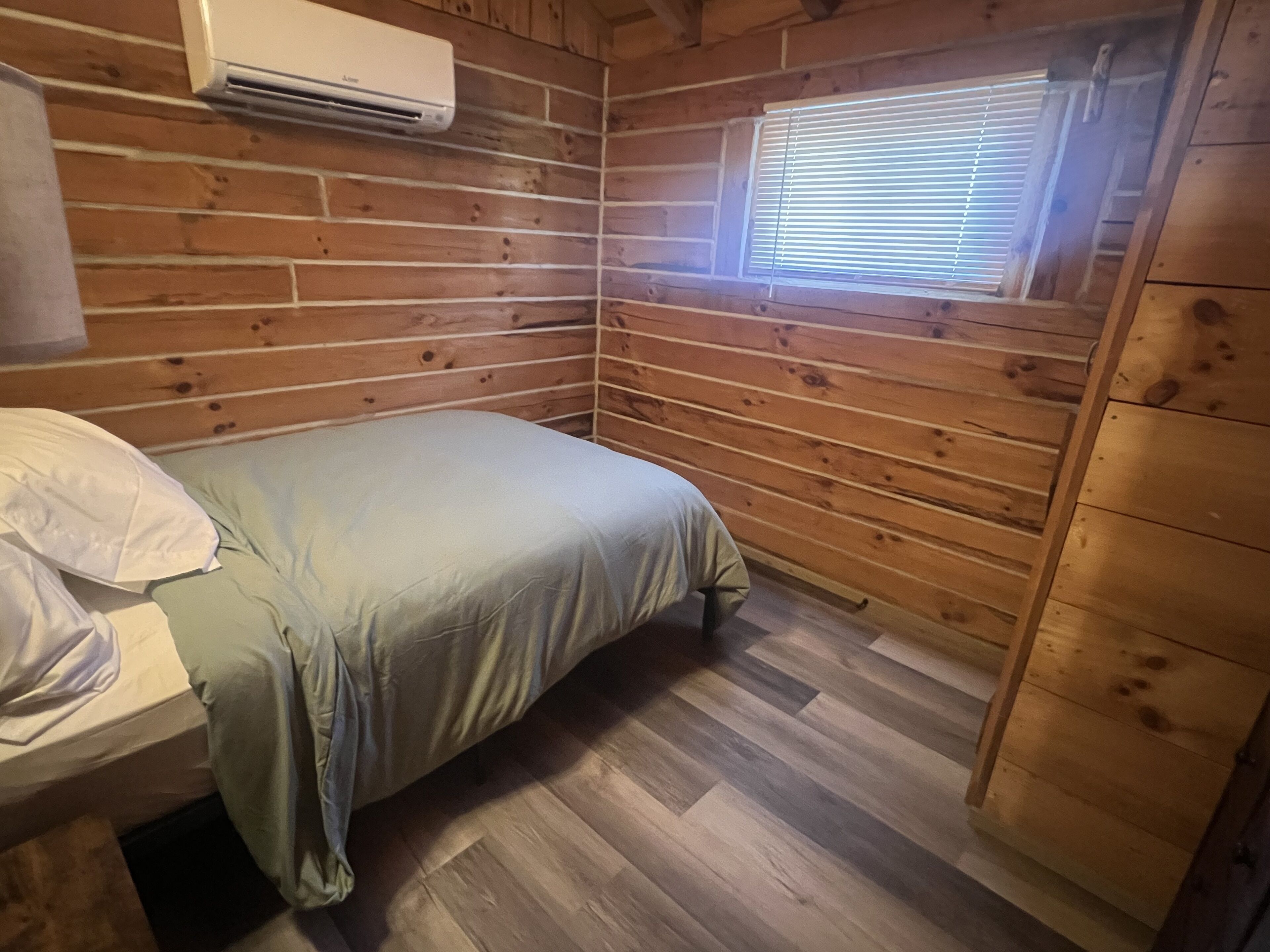 A small, cozy room with wooden walls, a single bed, a window with blinds, and an air conditioning unit.