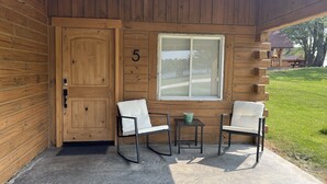 A wooden cabin with a number '5' on the door, two white chairs, a small table, and a window with blinds.