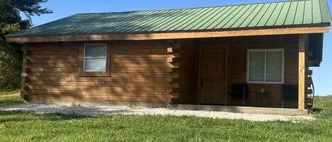 A wooden cabin with a green metal roof, a small window, and a door leading to a porch.
