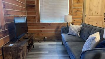 Interno di una baita in legno con pavimento in legno, un divano blu scuro con cuscini a righe, un mobile porta TV in legno e una finestra con persiane.
