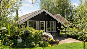 Une petite maison aux couleurs sombres, dotée d’un toit en pente, de grandes fenêtres et d’un jardin agrémenté de diverses plantes et d’une structure en bois.