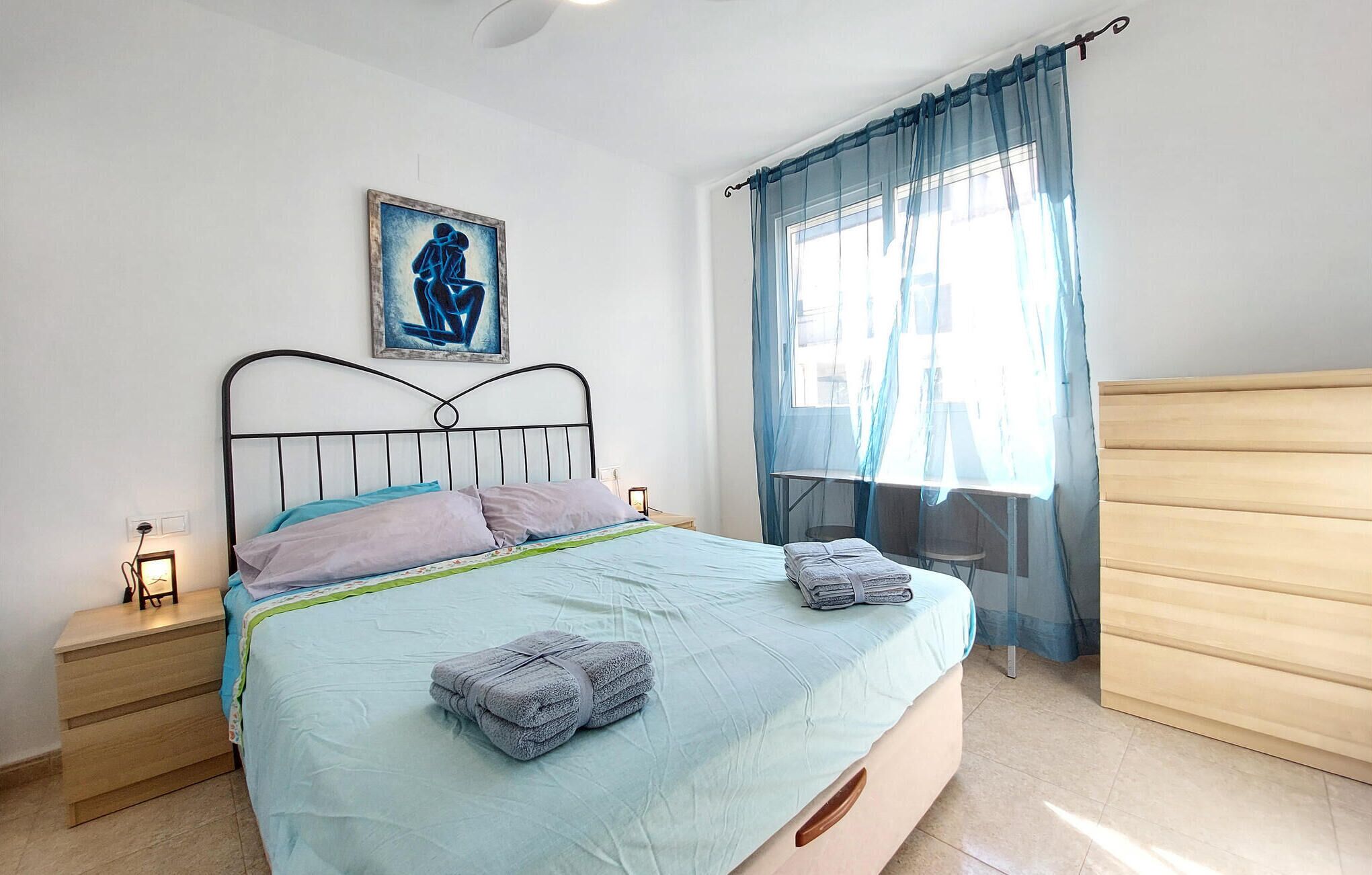 Een slaapkamer met een bed, een nachtkastje, een raam met gordijnen, een houten dressoir en een schilderij aan de muur.