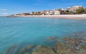 Una zona costera con aguas turquesas cristalinas, terreno submarino rocoso y una hilera de edificios a lo largo de la orilla.