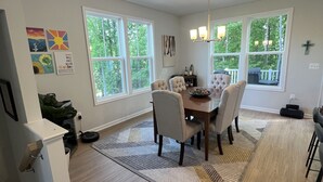 A dining area with a wooden table, six chairs, and a chandelier. There are large windows with a view of greenery outside.