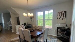 A spacious dining area with a wooden table and cushioned chairs, a chandelier, and a view of greenery through large windows.