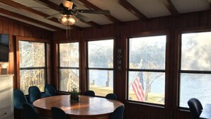 A dining area with a wooden table and chairs, large windows with a view of a body of water and trees, and a ceiling fan. - Riverfront ready for Summer Fun (Moss Point)