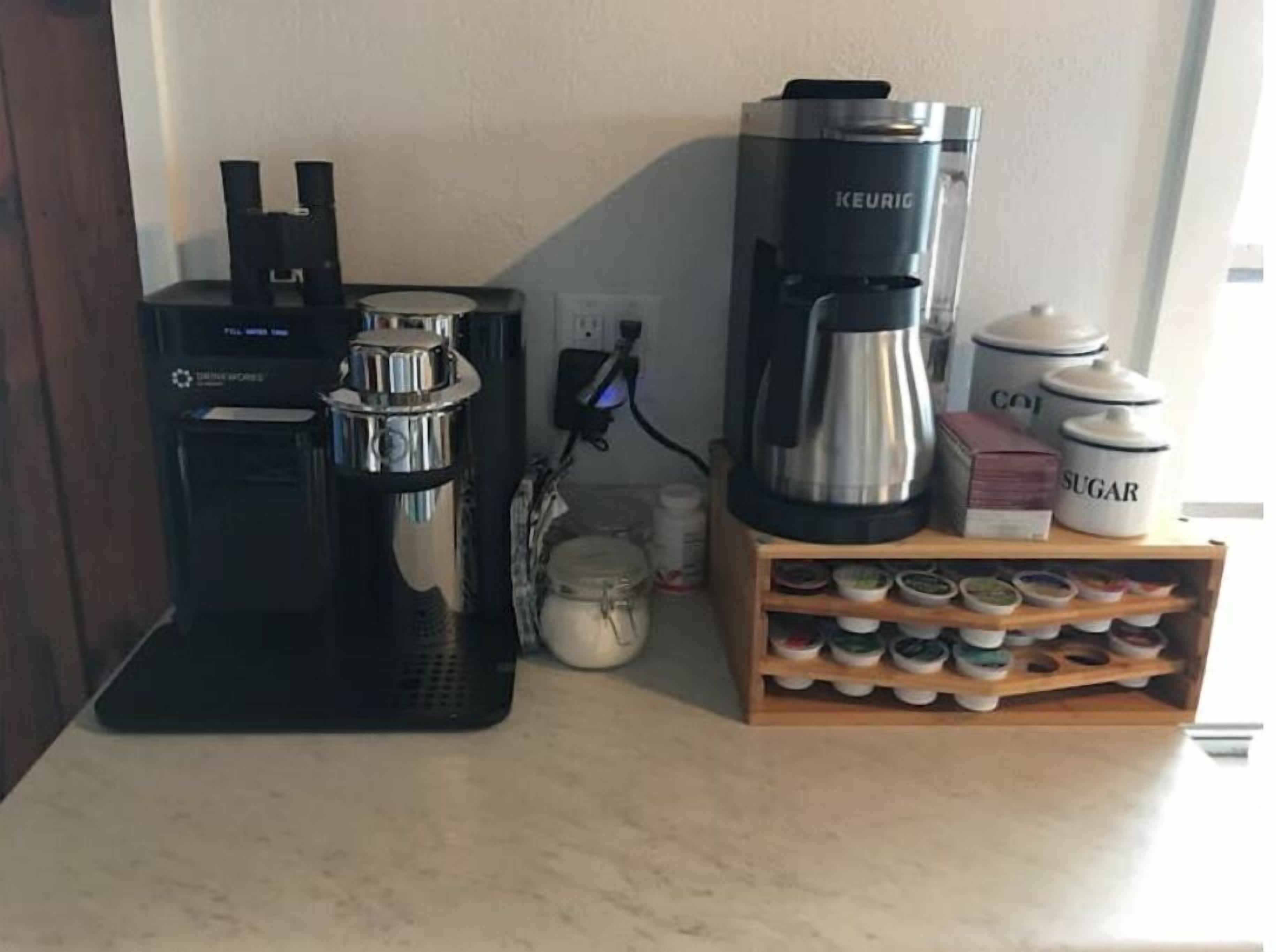 Une machine à café, un distributeur de dosettes de café Keurig, ainsi que des récipients à sucre et à crème, sont posés sur un plan de travail de la cuisine.