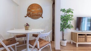 A modern living room with a round white table, four white chairs with brown cushions, a wooden sign above the table, a potted plant, and a TV on a wooden cabinet. - El Rincón de Cañadío · Charming Apartment in the Heart of Santander (Santander)