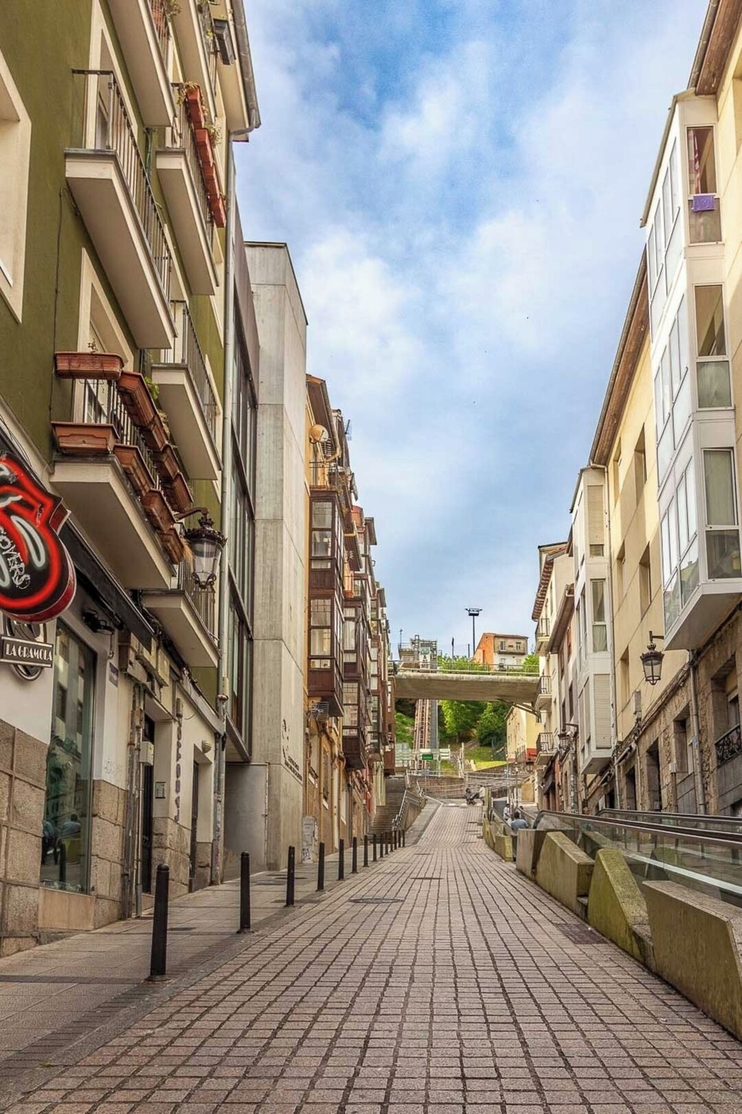 A narrow, paved street flanked by multi-story buildings with balconies, a pedestrian walkway, and a clear sky.