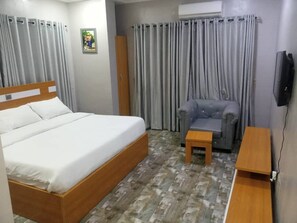 A hotel room with a wooden bed, grey curtains, a grey armchair, a small wooden table, and a TV on the wall. - Camas Hotel & Suite Asero Abeokuta (Abeokuta)