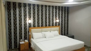 A neatly made bed in a hotel room with a patterned wallpaper, a bedside table with a phone, and a small nightstand. - Camas Hotel & Suite Asero Abeokuta (Abeokuta)