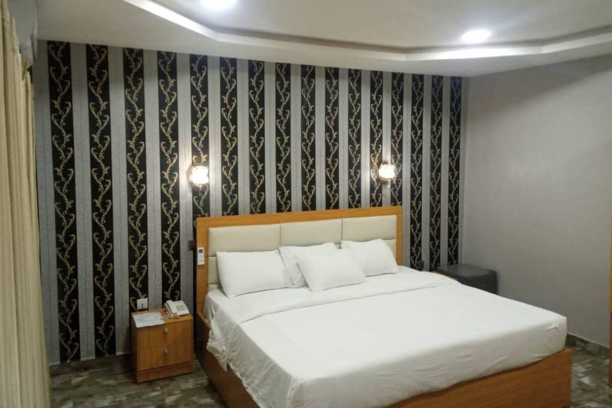 A neatly made bed in a hotel room with a patterned wallpaper, a bedside table with a phone, and a small nightstand.