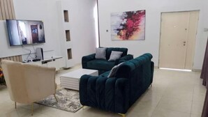 A living room with a beige armchair, a dark green sofa, a flat-screen TV mounted on the wall, and a door with a colorful abstract painting above it. - "Luxury 4-Bedroom Duplex in Lekki Lagos - Your Perfect Getaway (Lagos)