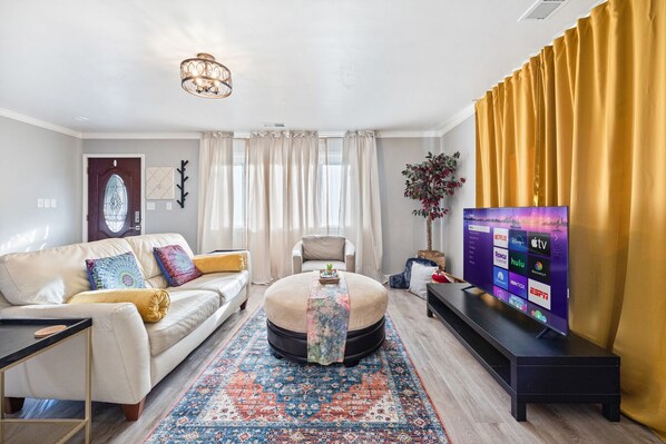 A modern living room with a beige sofa, a round ottoman, a coffee table, and a flat-screen TV displaying streaming services.