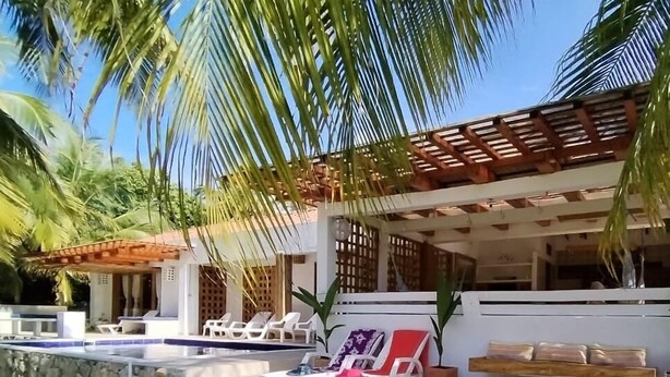 Un área junto a la piscina con sillones de descanso, una pérgola de madera y un muro de piedra.