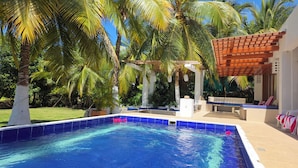 Una piscina con una pérgola y sillones de descanso, rodeada de palmeras y un césped.