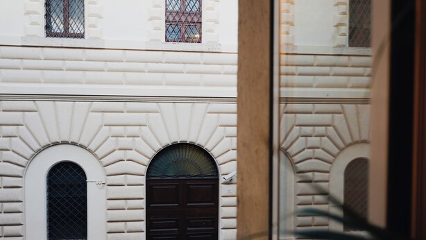 A building facade with arched windows and a central door, viewed through a window with a plant in the foreground.
