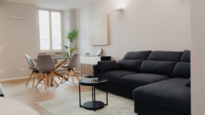 A modern living room with a dark grey sectional sofa, a round coffee table, and a dining area with a glass table and grey chairs.