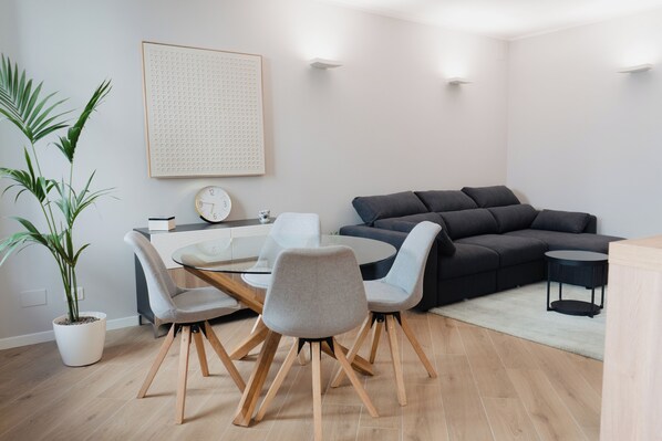 Uma sala de estar moderna com uma mesa de jantar redonda, cadeiras cinzas, um sofá cinza escuro, uma mesinha lateral e um vaso com planta.