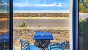 Un balcon avec un mur en pierre, une table et deux chaises, donnant sur une plage et l’océan.