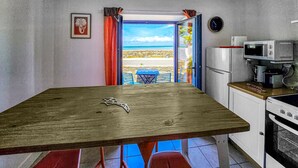 Une petite cuisine avec une table en bois, un réfrigérateur, un four à micro-ondes et une cuisinière. Il y a une fenêtre avec des rideaux et une vue sur l’extérieur.