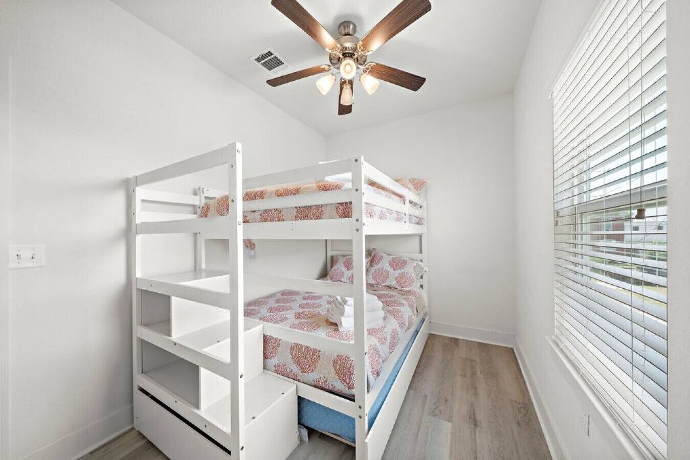 A bunk bed room with white walls, a ceiling fan, and a window with blinds.