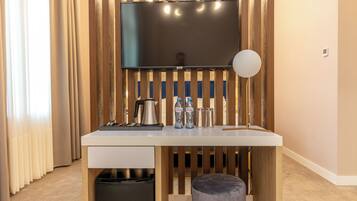 Ein modernes Hotelzimmer mit weißem Schreibtisch, einem flachbildfernseher an einer Holzvertäfelung, einer schwarzen Mülltonne und einem grauen Pouf.