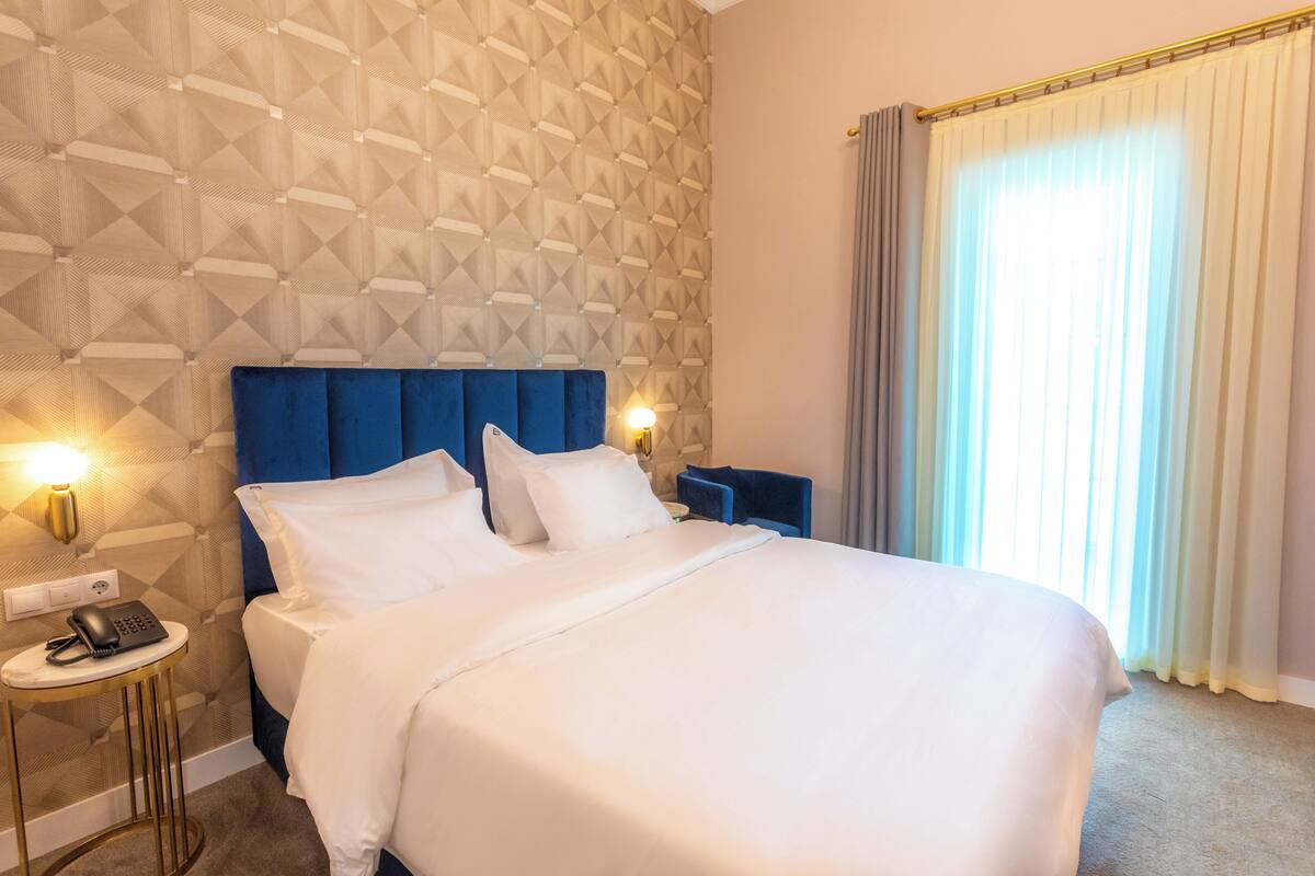 Habitación de hotel con una cama grande, cabecera, dos mesitas de noche, teléfono y ventana con cortinas.