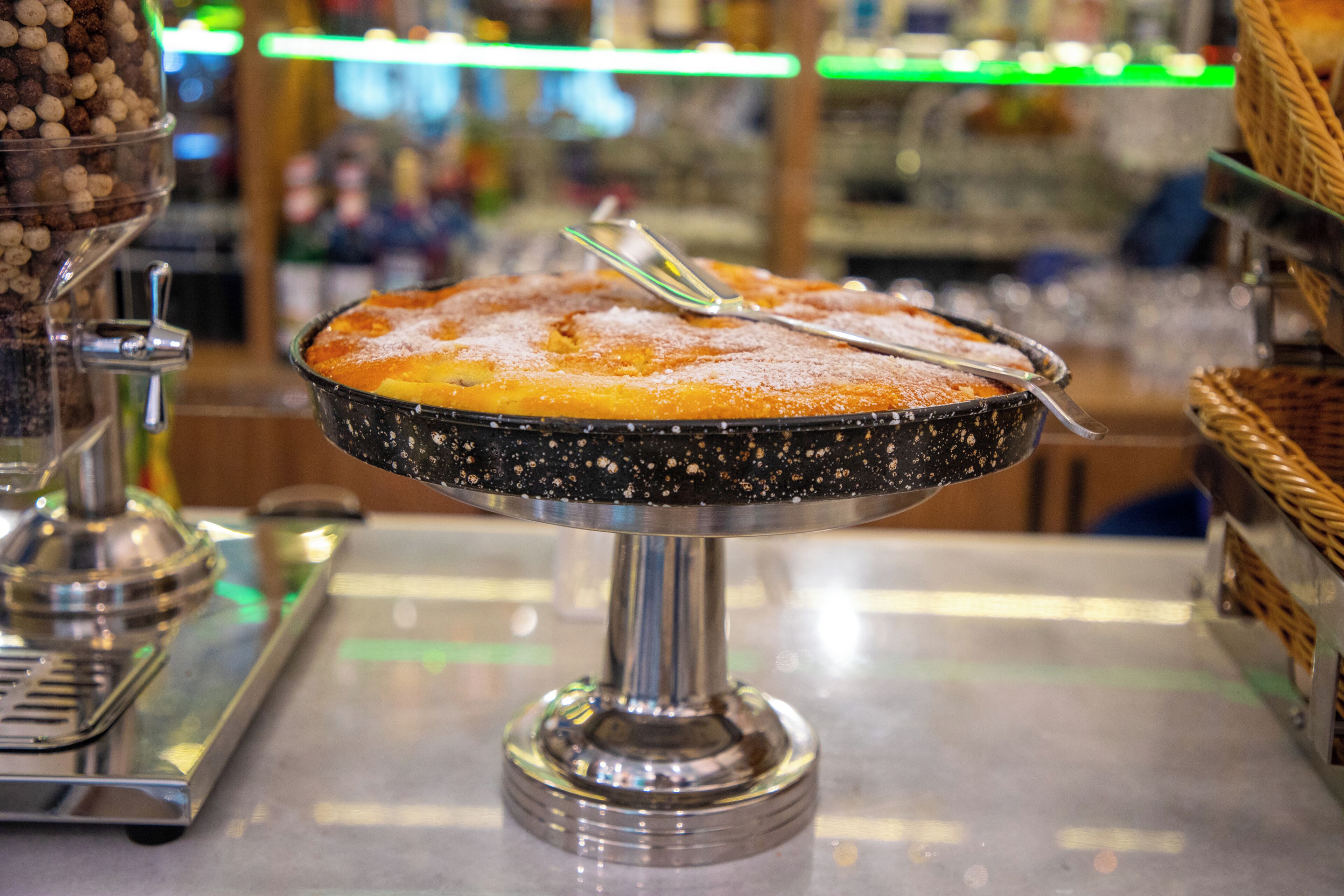 A round cake on a metal stand with a black speckled base, dusted with powdered sugar, and a serving spoon on top.