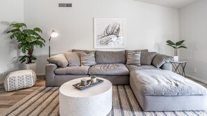 Een moderne woonkamer met een grijze hoekbank, een ronde salontafel en twee planten in potten.