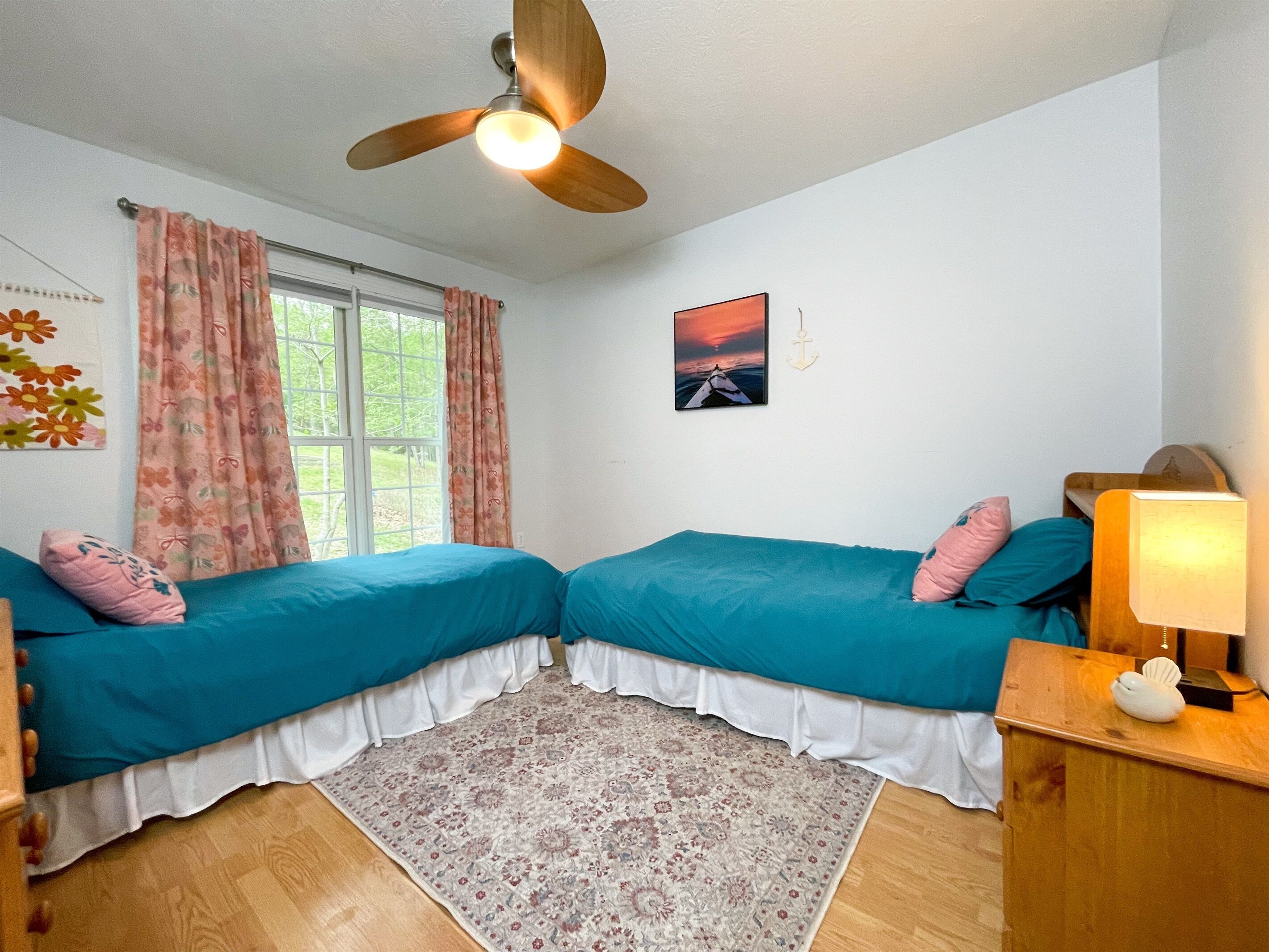 Une chambre avec deux lits, un ventilateur de plafond, une fenêtre avec des rideaux, un tapis et un tableau accroché au mur.